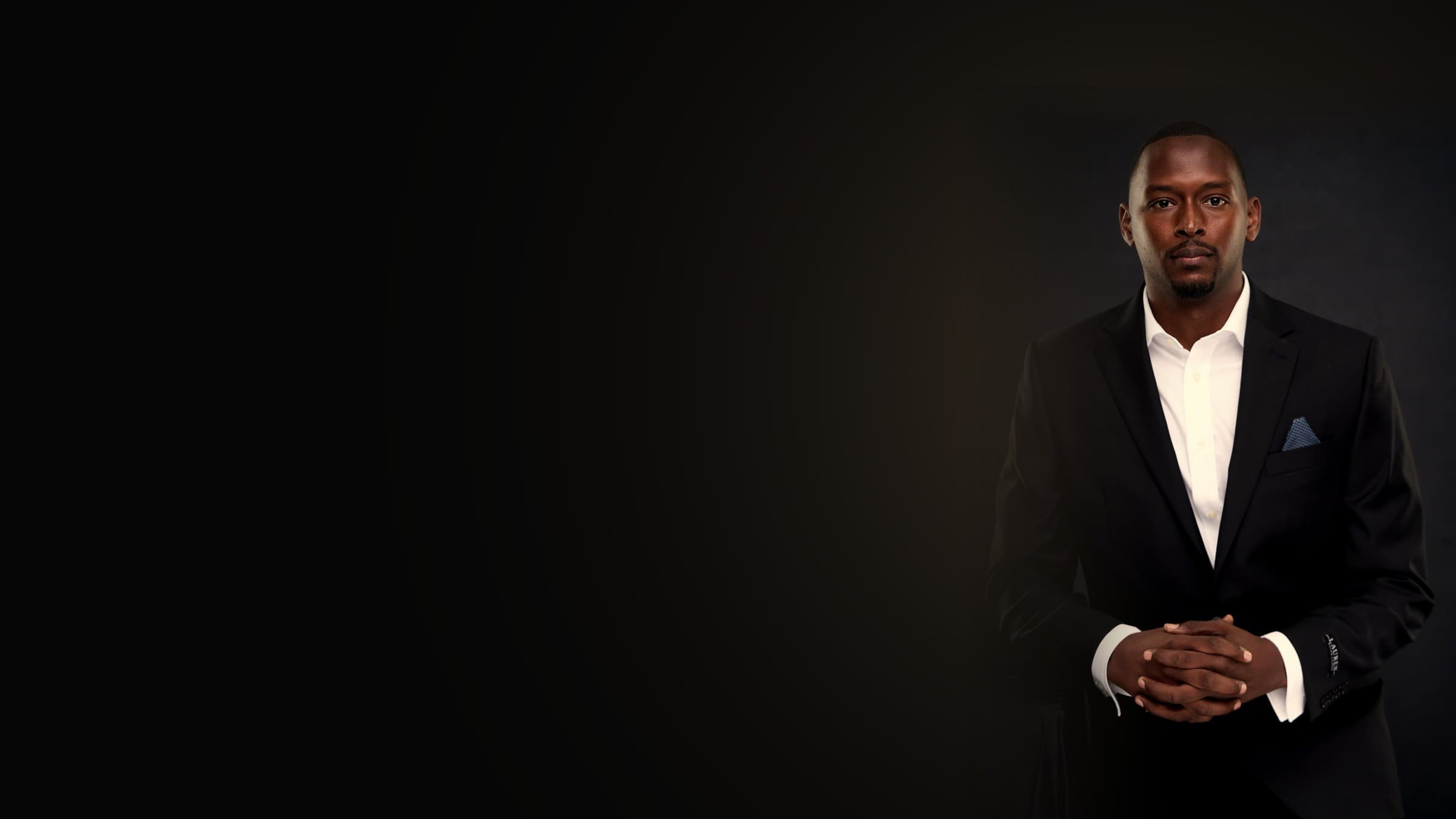Osama Mansour in a dark suit, hands clasped, against a near-black studio backdrop.